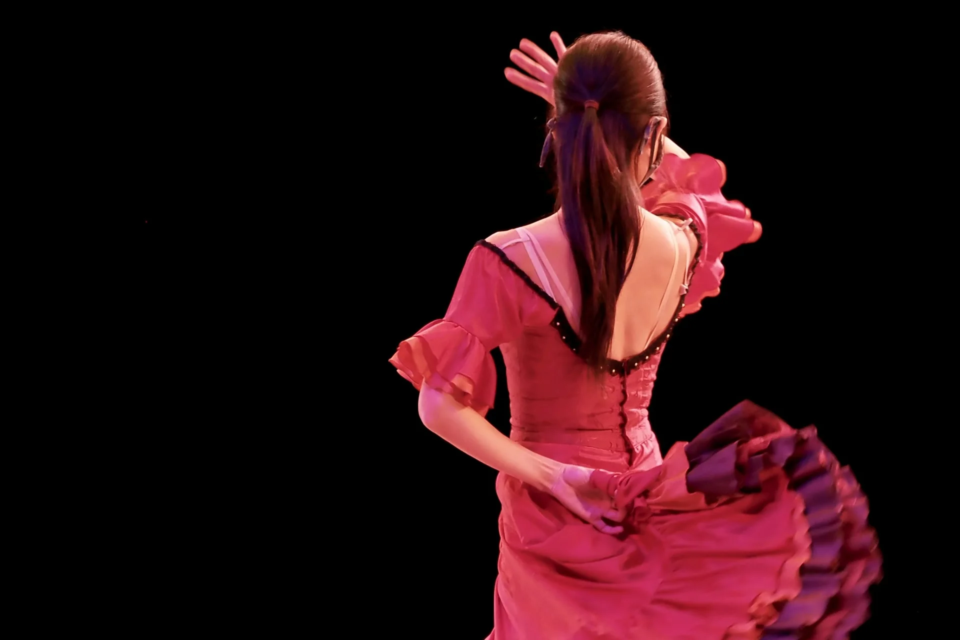 Flamenková tanečnice v půlobratu v růžových volánových šatech na tmavém jevišti, sukně rozvlátá v pohybu (ilustrativní fotografie flamenkového představení, nejde o El Palacio Andaluz)
